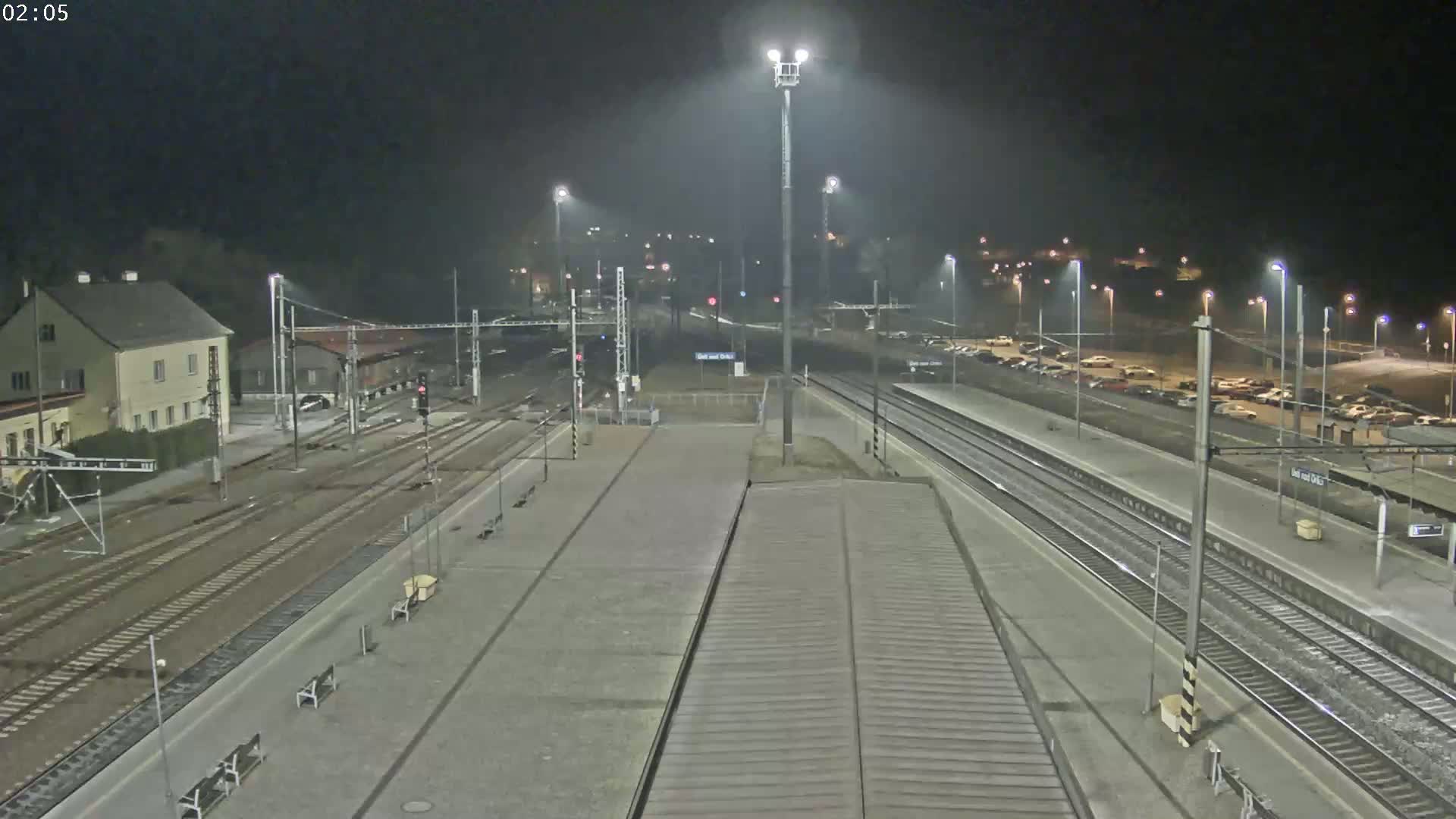 A nighttime view of a mostly empty train station with multiple tracks and a parking lot, under hazy or foggy conditions.