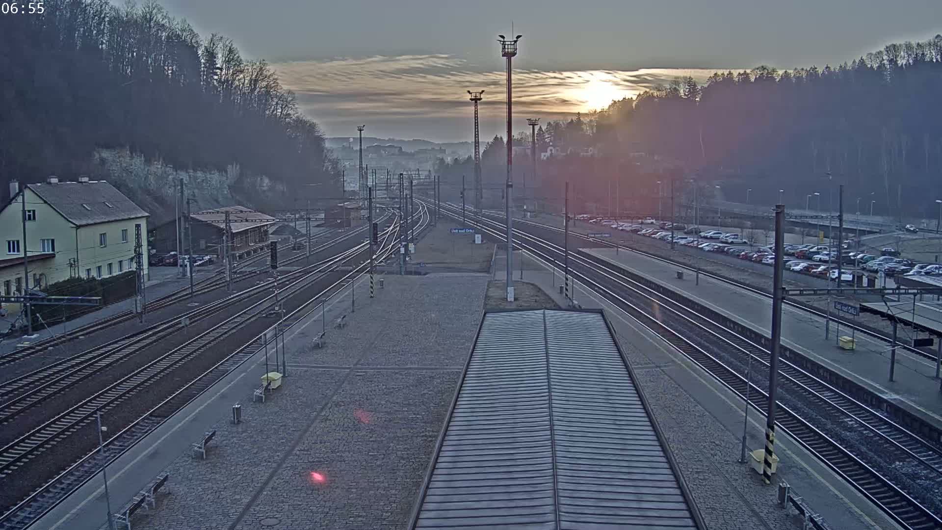 A high-angle, long shot of a train station at sunrise, showing multiple train tracks, a parking lot filled with cars, and buildings in the background.