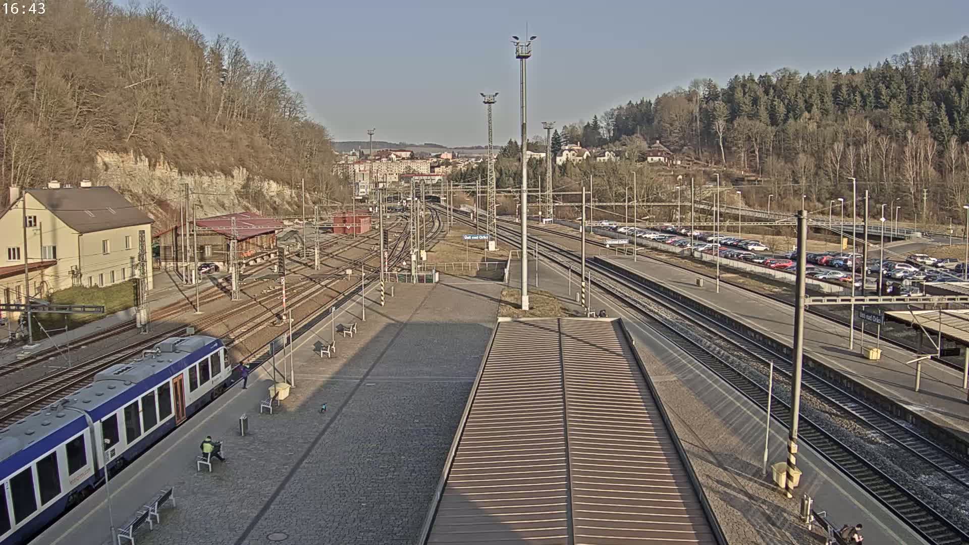 A train station on a sunny day with multiple train tracks, a passenger train at the platform, and a large parking lot filled with cars.