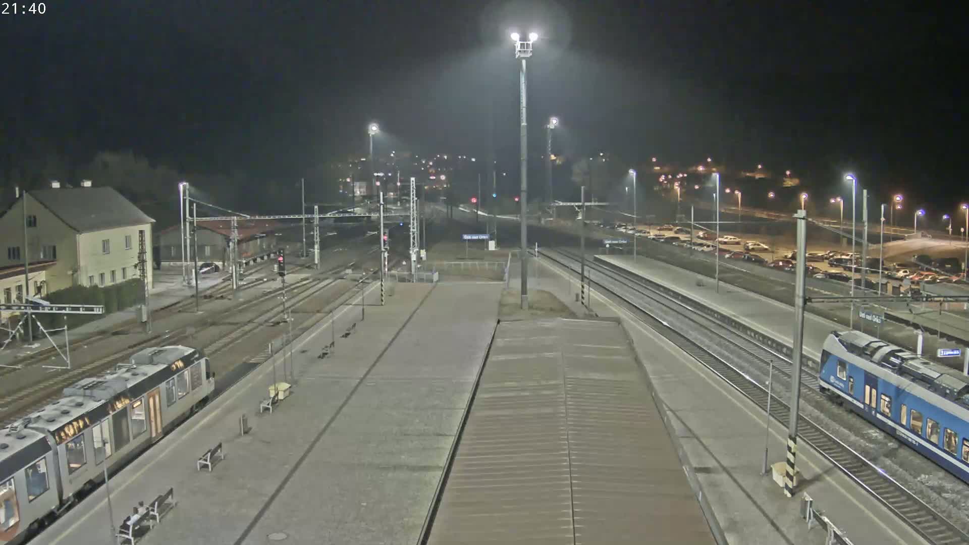 A nighttime view of a train station with two trains at the platform, under partly cloudy skies, and several cars parked in a nearby lot.