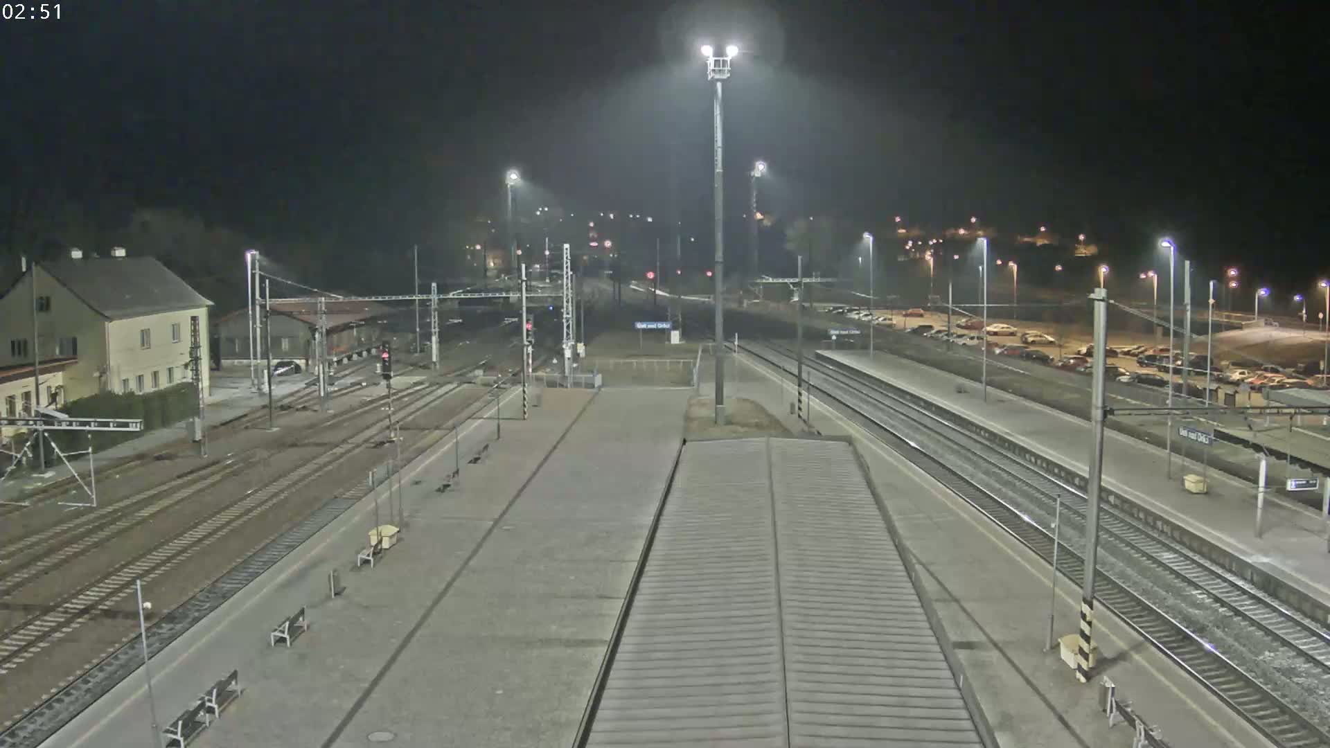 A nighttime view of a train station with multiple train tracks, platforms, and a parking lot, illuminated by tall lights.