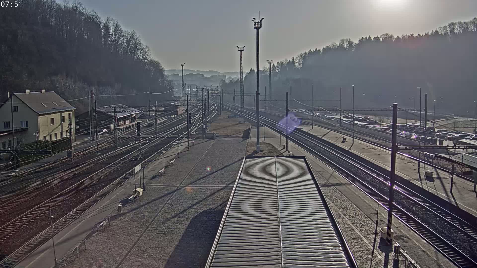 A high-angle, long shot of a train station on a sunny, hazy day, showing multiple train tracks, a building, and a parking lot filled with cars.