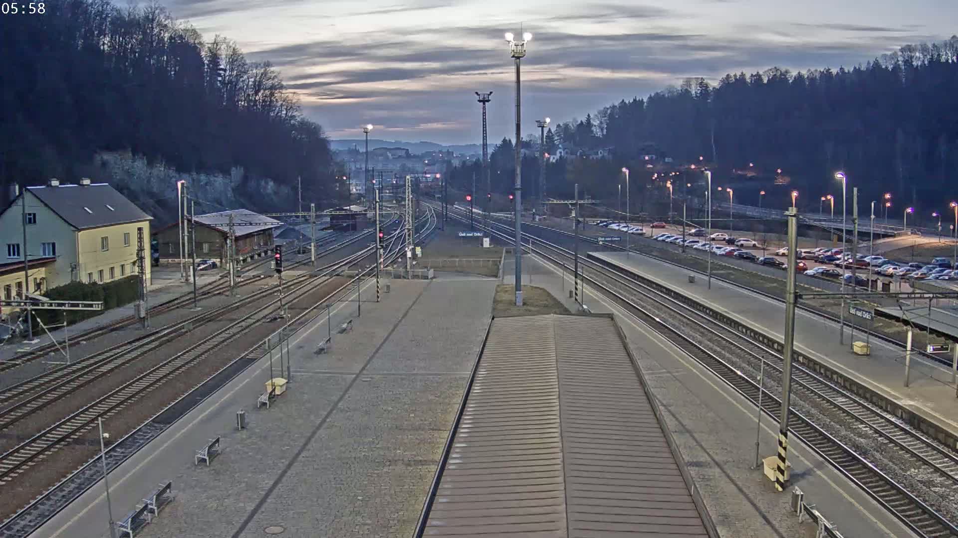 A train station with multiple tracks, a parking lot, and a building is visible under a dusky sky.