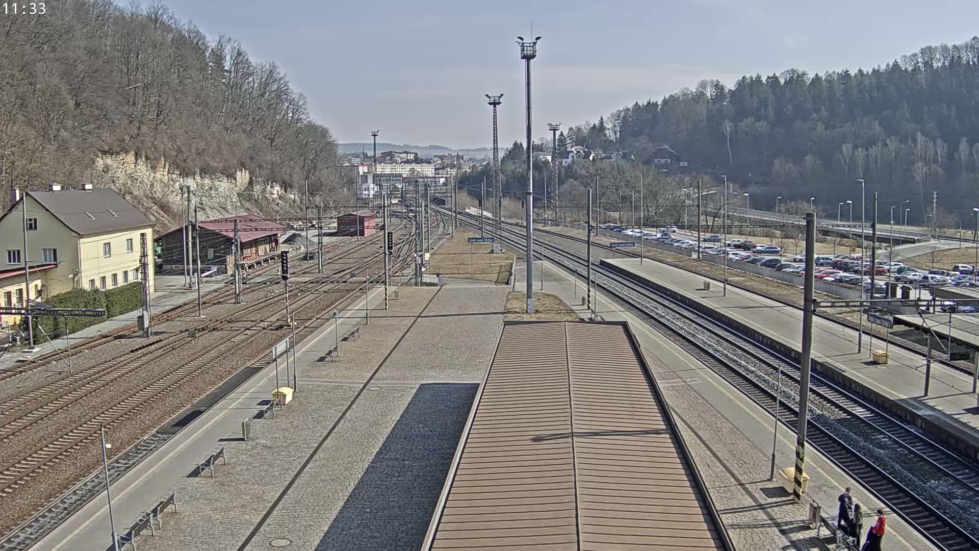 A sunny view of a train station with multiple train tracks, a platform, and a parking lot, surrounded by trees and buildings.