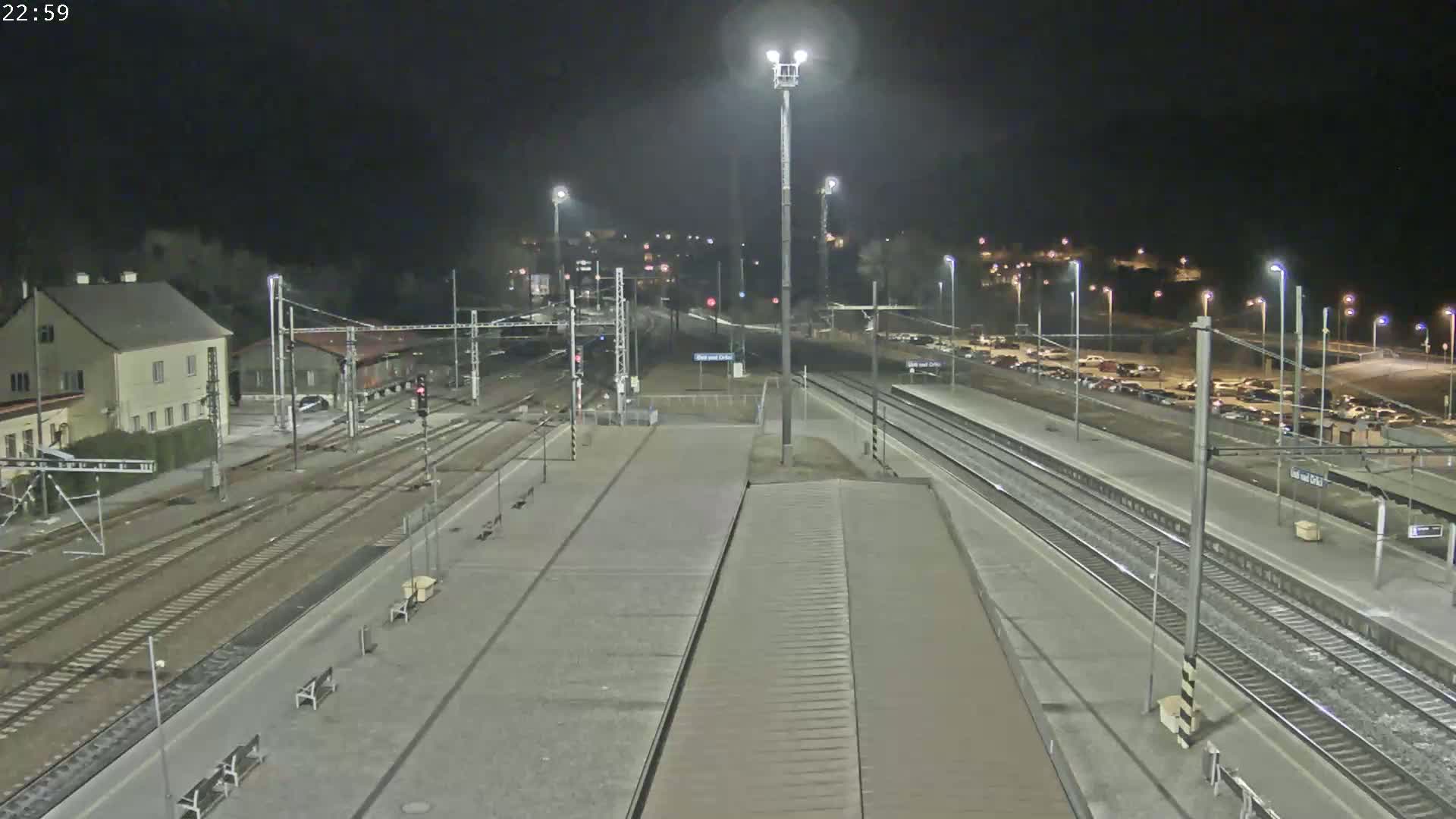 A nighttime view of a train station with multiple tracks, platforms, and a parking lot, illuminated by artificial lights.