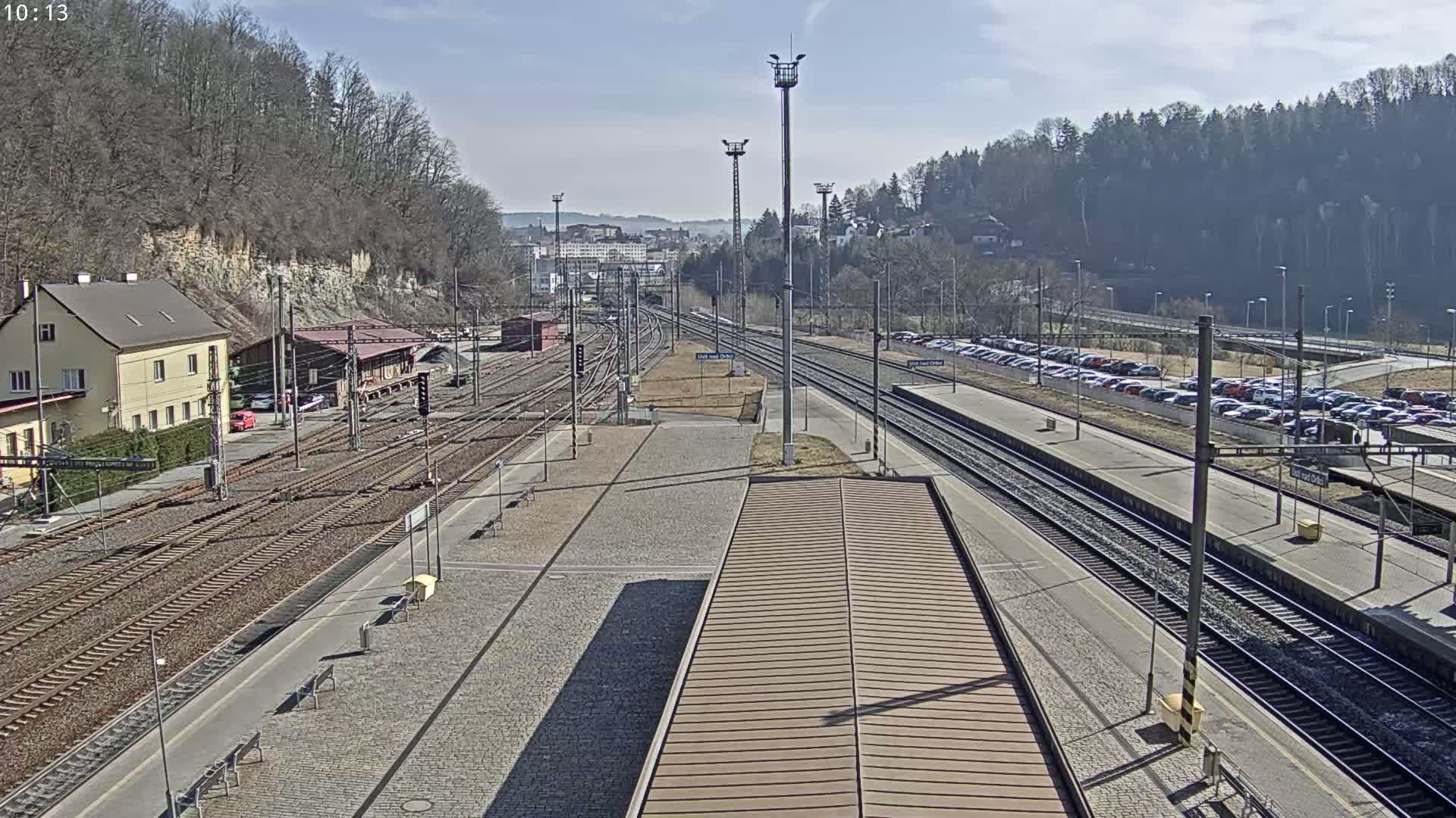A sunny view of a train station with multiple train tracks, a large parking lot, and a building situated on a hillside.