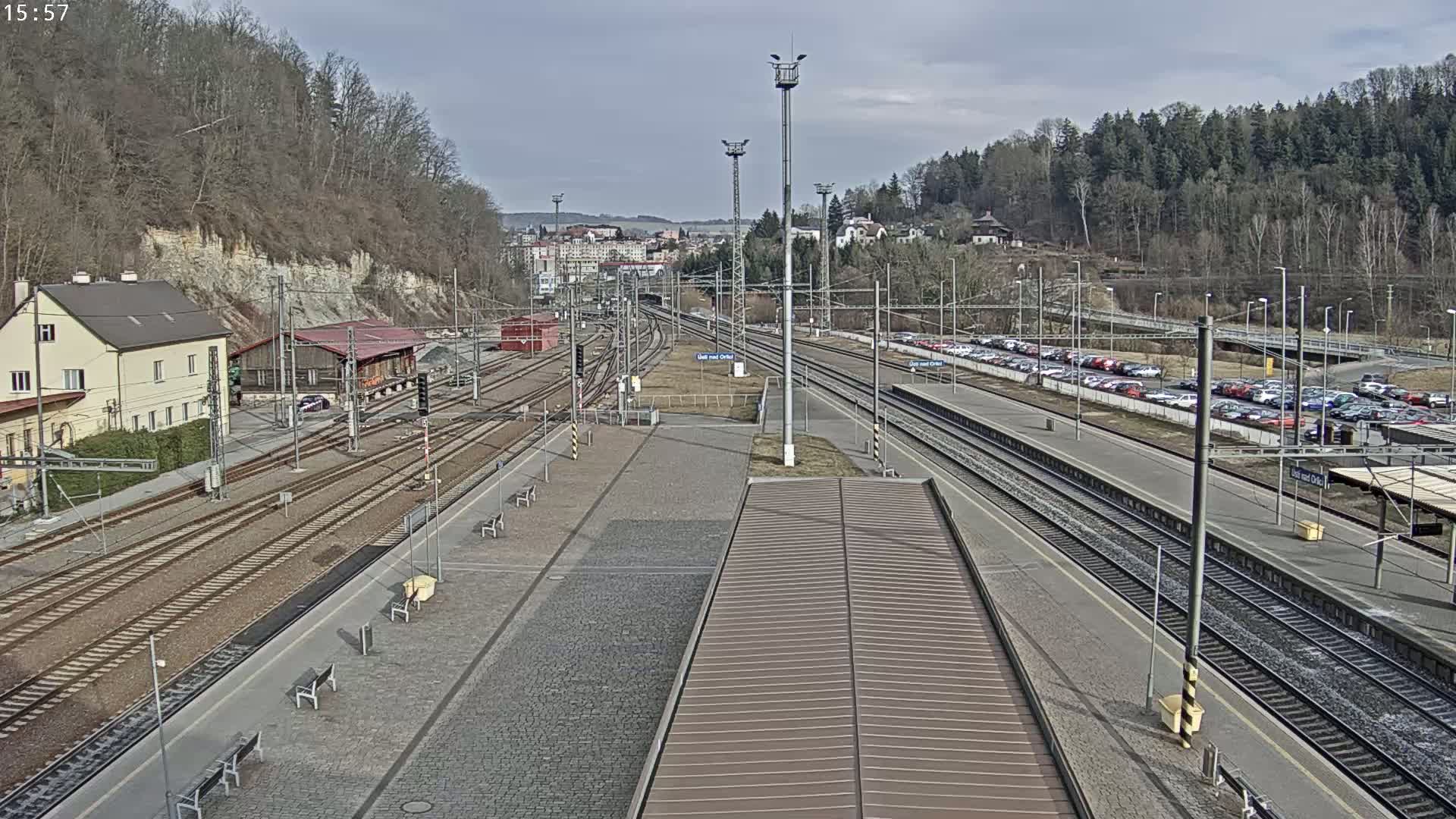 A train station with multiple tracks, a parking lot full of cars, and buildings nestled beside a hillside under a partly cloudy sky.