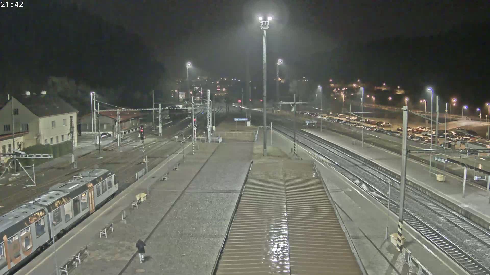A nighttime view of a train station with a train at the platform, numerous tracks, and a large parking lot with cars under artificial lighting.