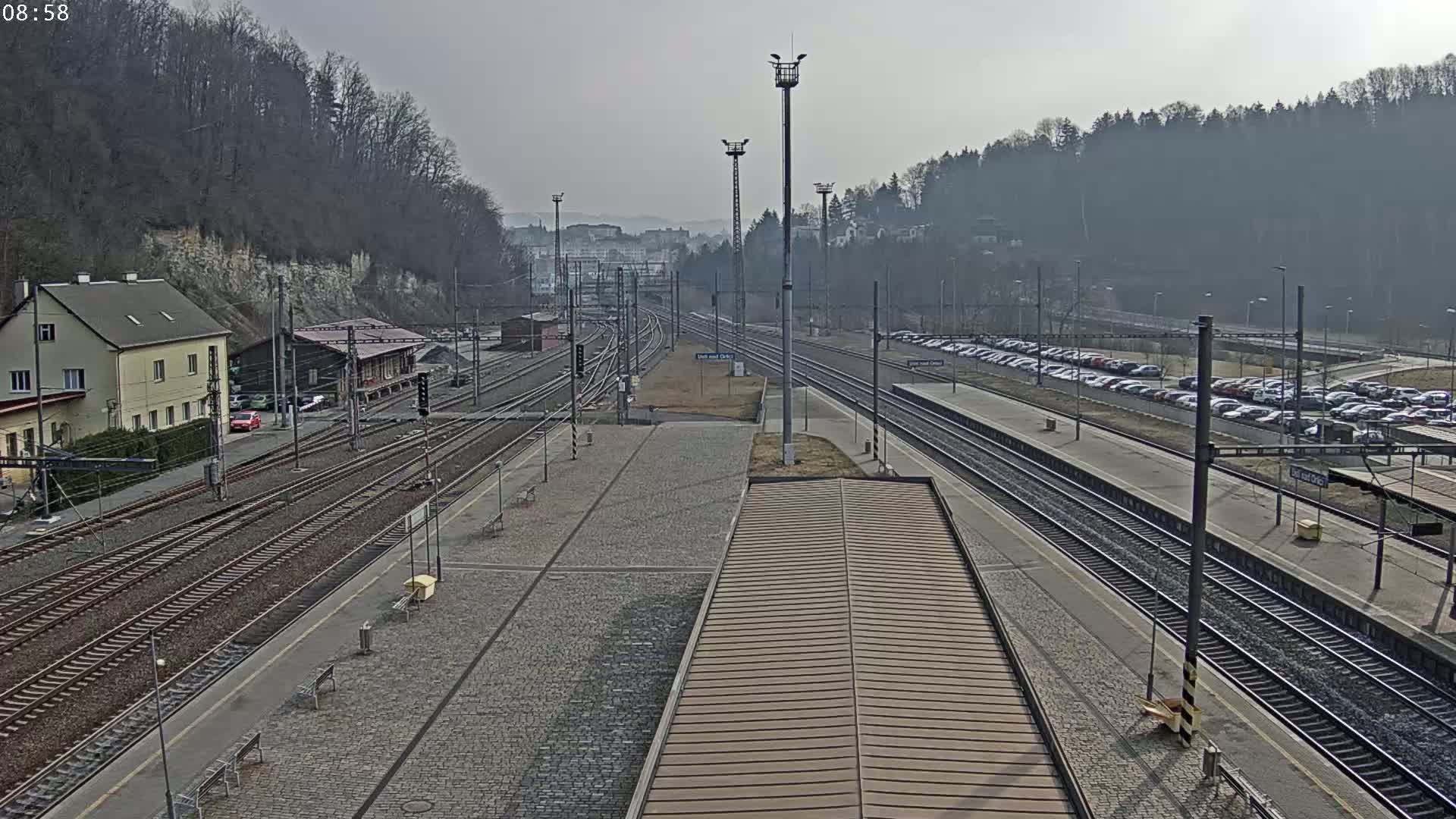 A train station with multiple train tracks, a building, a parking lot filled with cars, and a hillside covered in leafless trees under an overcast sky.