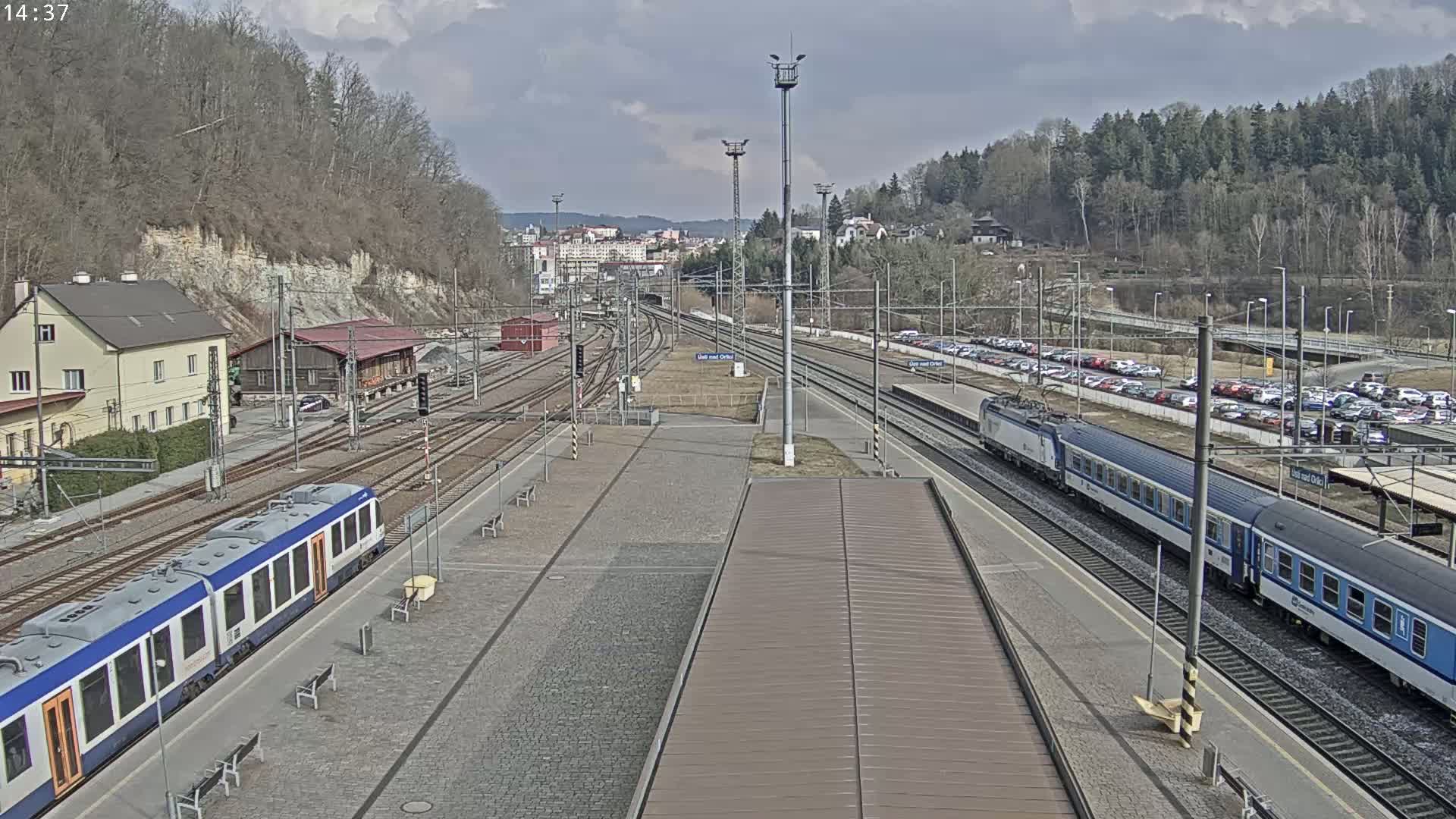 A train station with two trains on the tracks, under a partly cloudy sky, surrounded by buildings and a hillside with leafless trees.