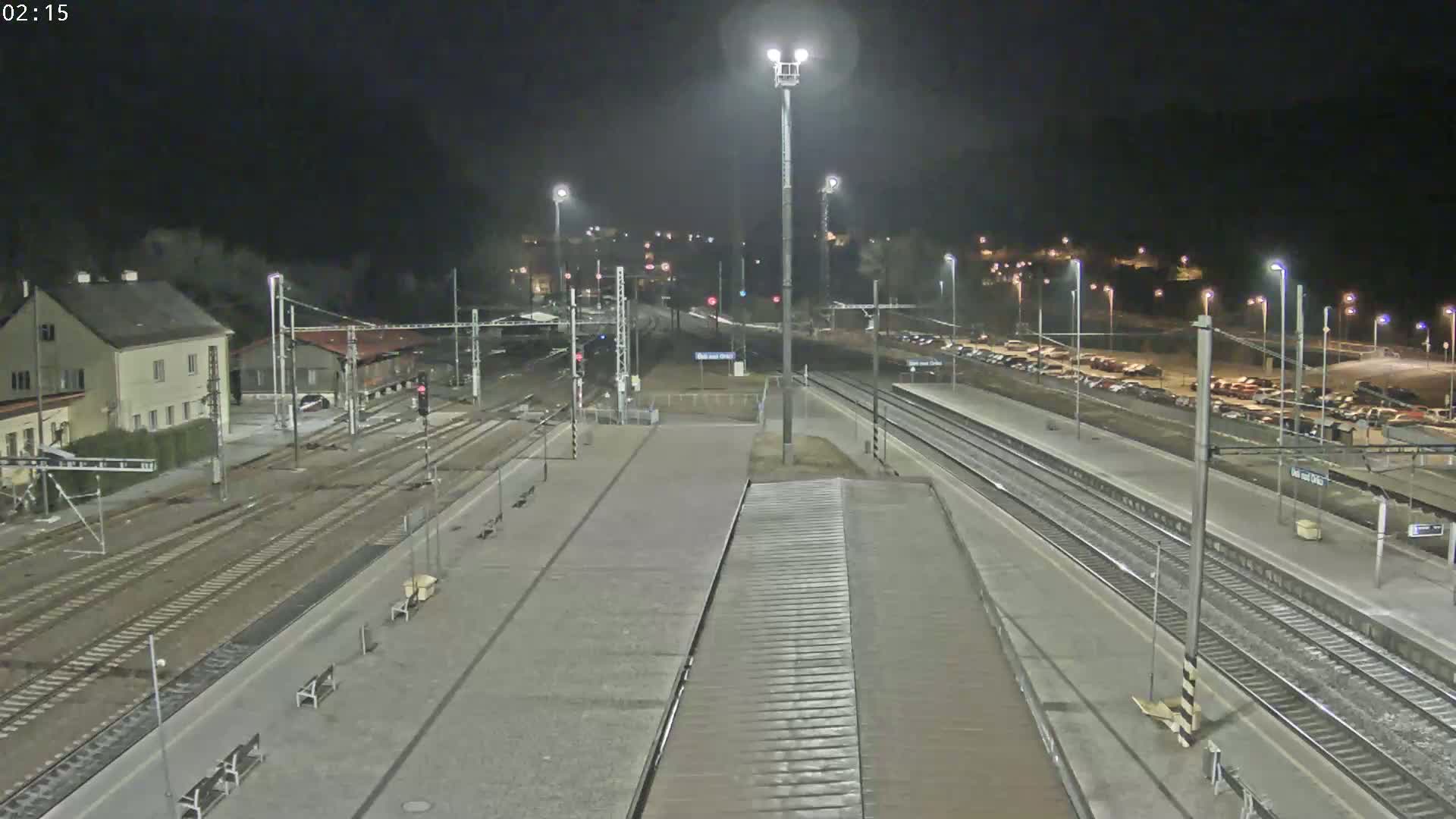 A nighttime view of a train station with multiple train tracks, platforms, and a parking lot, under clear skies.