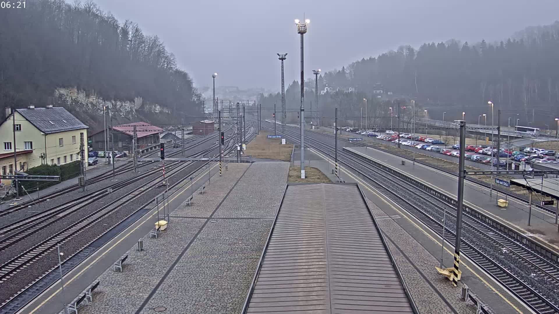 A train station with multiple tracks, a parking lot filled with cars, and buildings, all under a foggy sky.