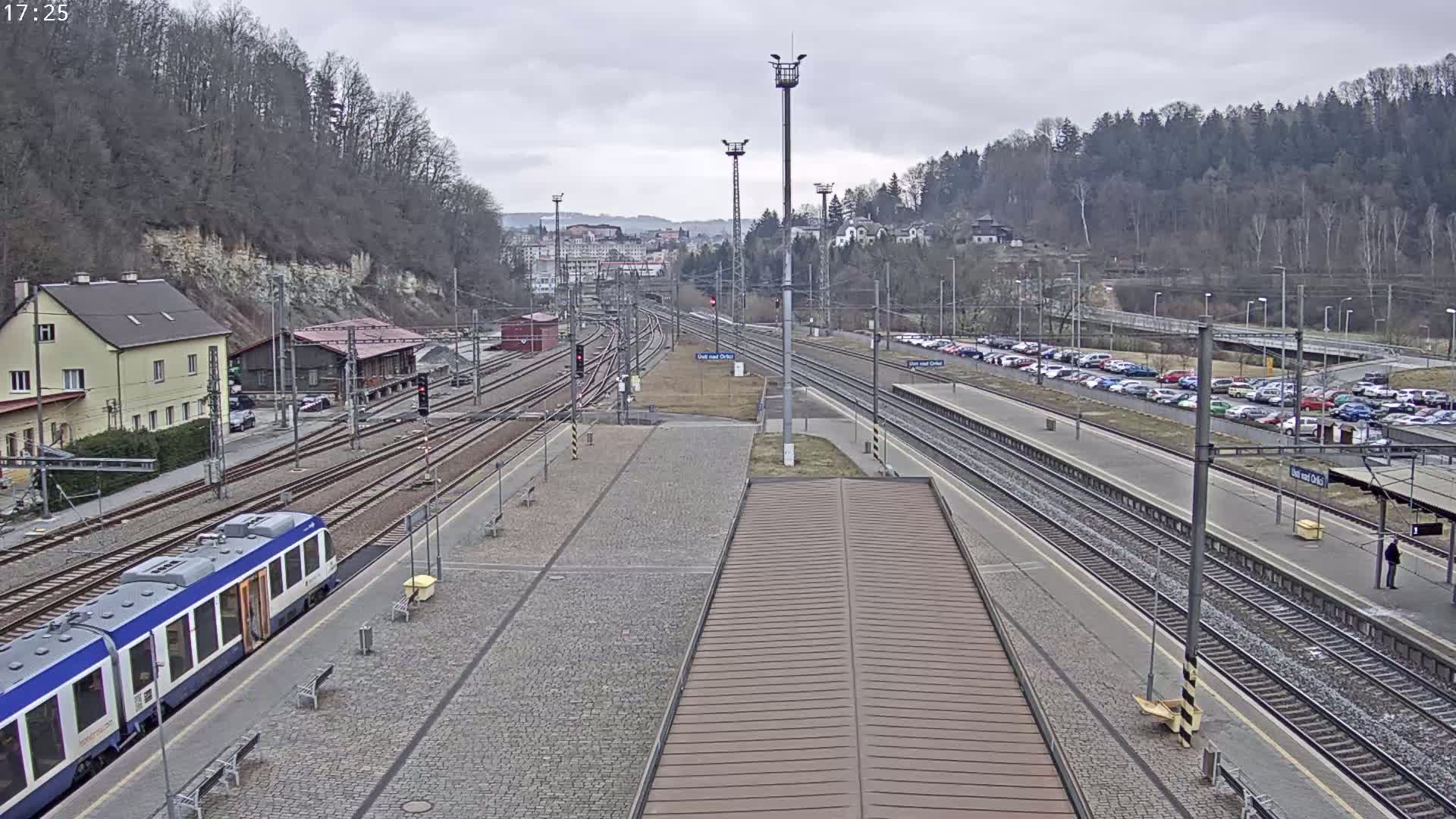 A train station with multiple train tracks, a train, and a large parking lot is viewed on an overcast day.