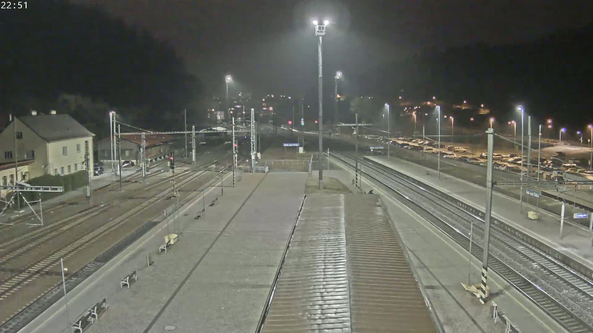 A nighttime view of a train station with multiple tracks, a parking lot filled with cars, and a building in the background.