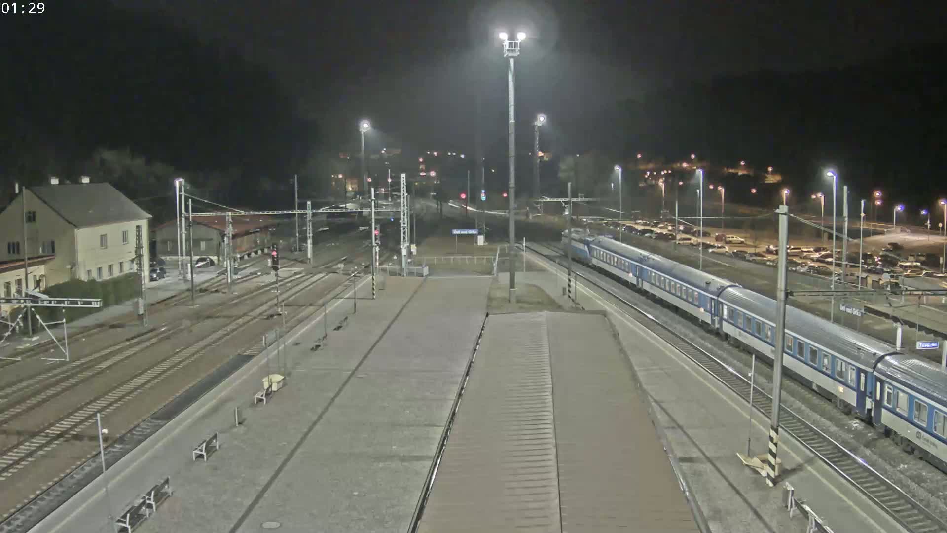 A long passenger train is at a nighttime train station with multiple tracks and a parking lot visible in the background under a dark sky.