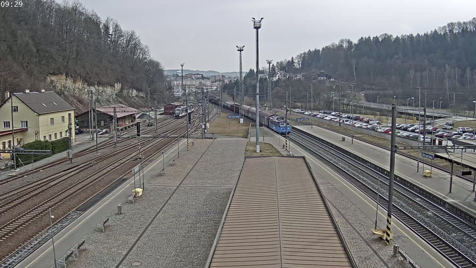 A train station with multiple tracks, a long freight train, and a large parking lot is visible on an overcast day.