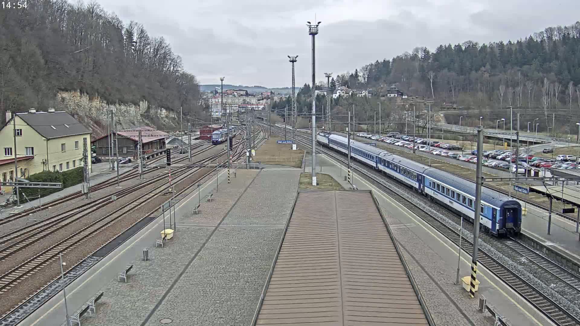 A train station on an overcast day with two trains on tracks, surrounded by buildings and a parking lot with numerous cars.
