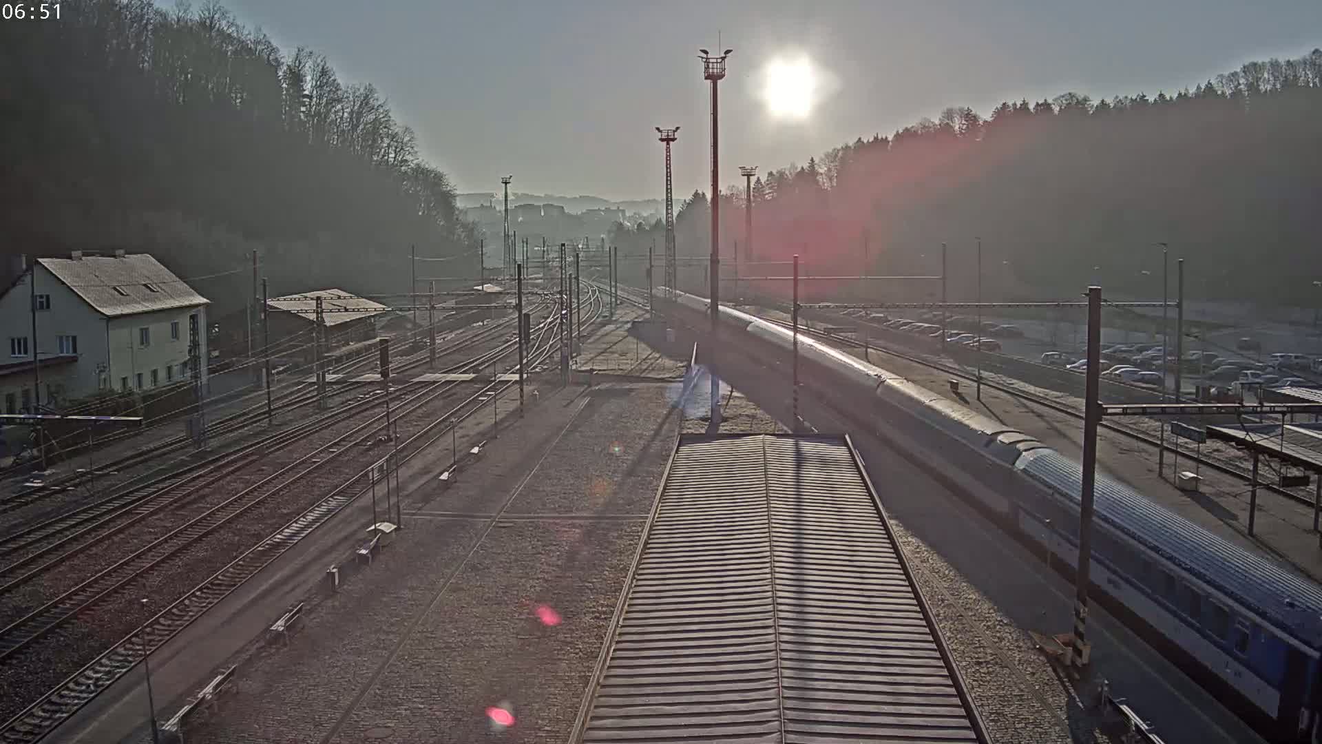 A long train sits at a station platform on a sunny, hazy morning, with multiple train tracks and a parking lot visible.