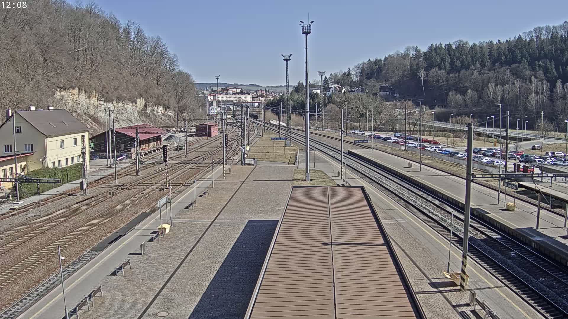 A train station with multiple tracks and a large parking lot is seen on a sunny day, surrounded by trees and hills.