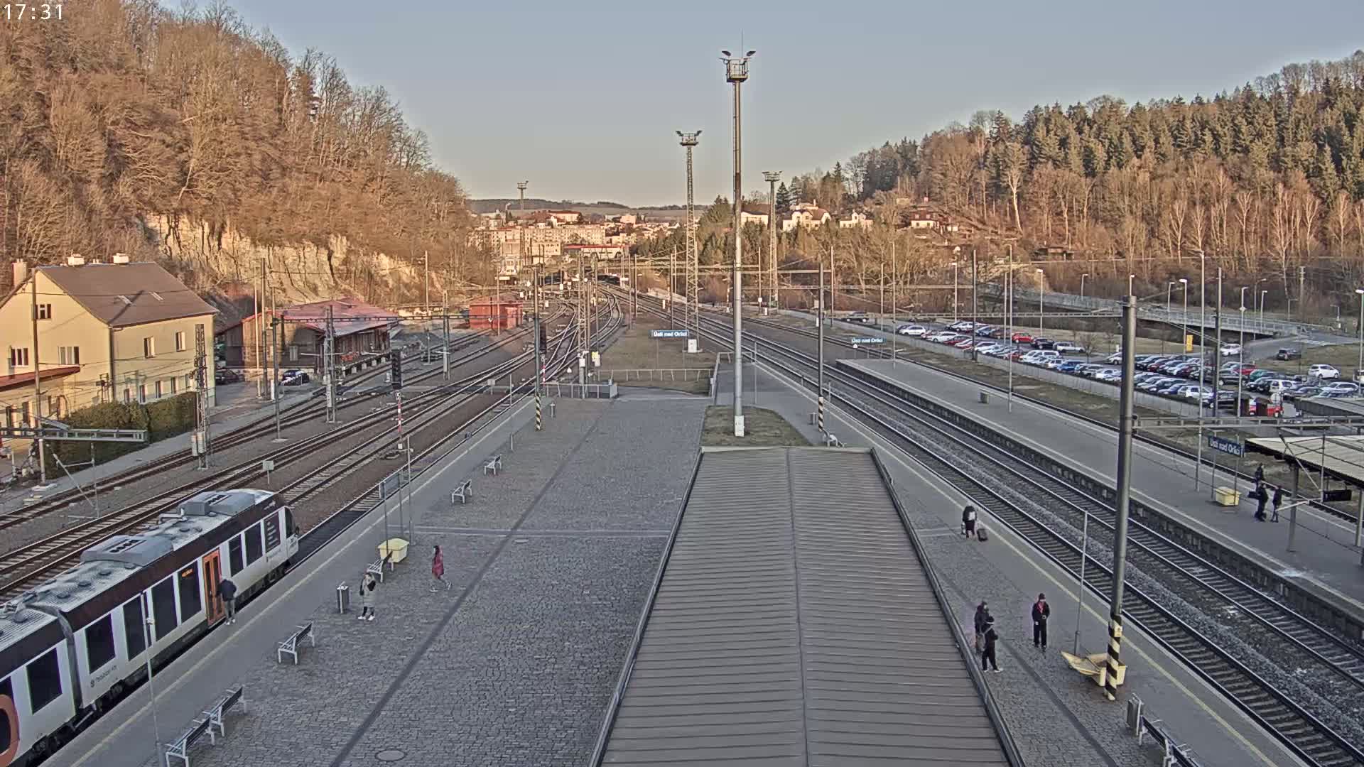A train station with multiple tracks, a train at the platform, a parking lot, and a few people walking around on a clear, sunny day.