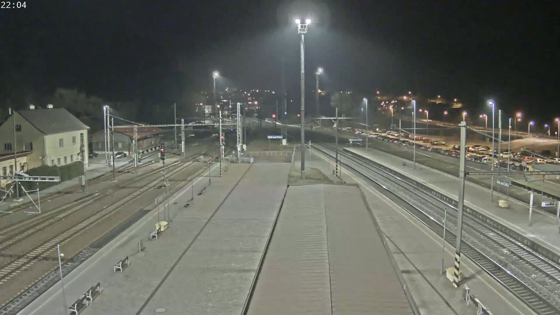 A nighttime view of a train station with multiple train tracks, platforms, and a parking lot filled with cars under bright artificial lighting.