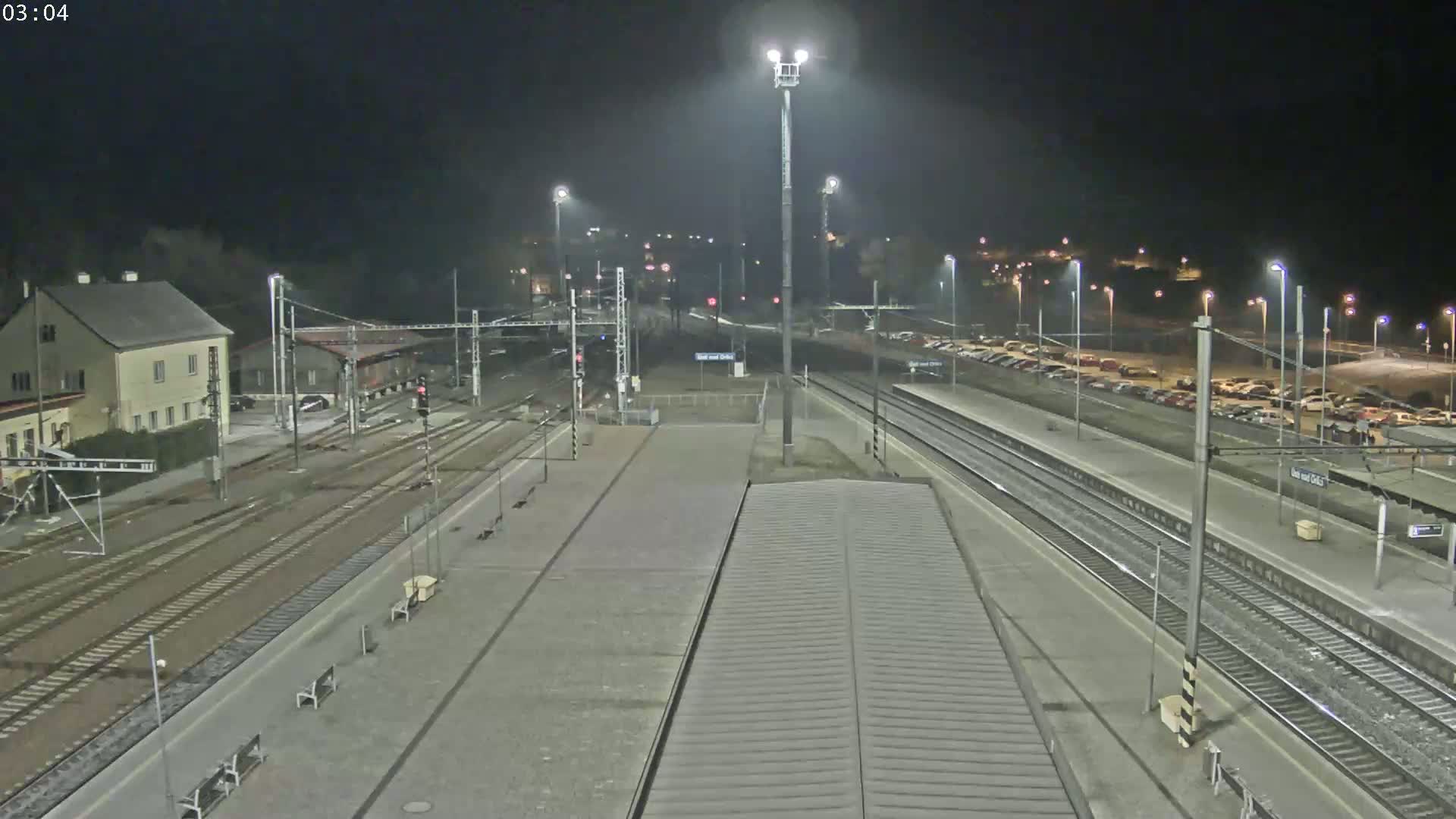 A nighttime view of a train station with multiple tracks, a platform, a parking lot, and a building, under dark and clear skies.