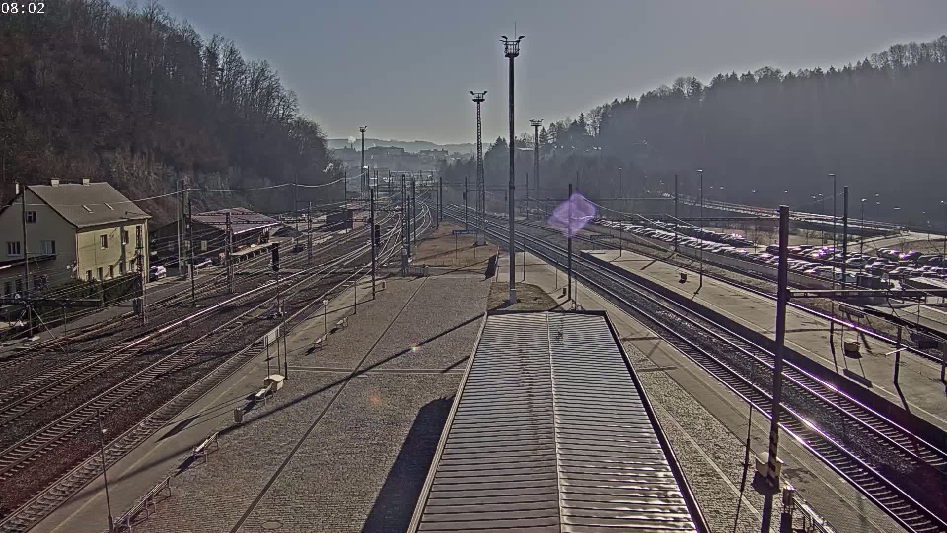 A sunny day at a train station with many tracks, a building, and a large parking lot filled with cars, all set against a backdrop of trees and hills.