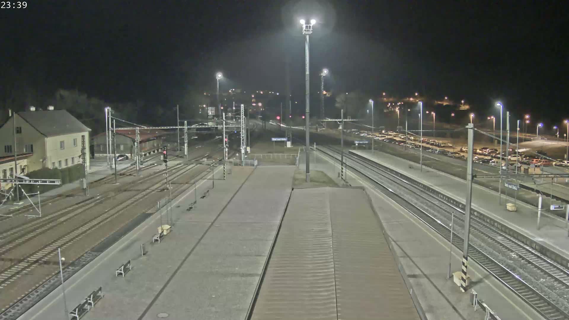 A nighttime view of a train station with multiple tracks, platforms, and a parking lot filled with cars under dark, clear skies.