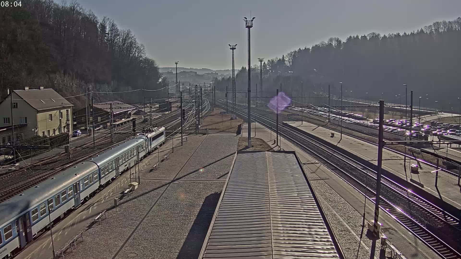 A passenger train is arriving at a train station on a sunny day, with multiple train tracks and a large parking lot visible.