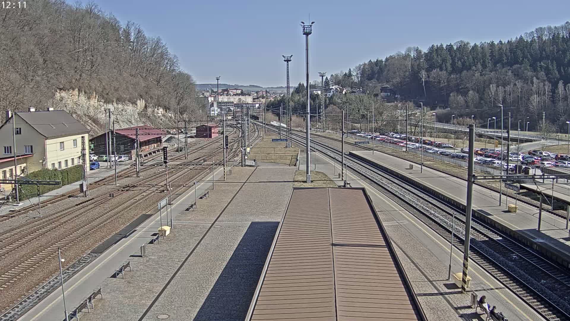 A train station on a sunny day with multiple train tracks, a large parking lot filled with cars, and buildings surrounding the area.