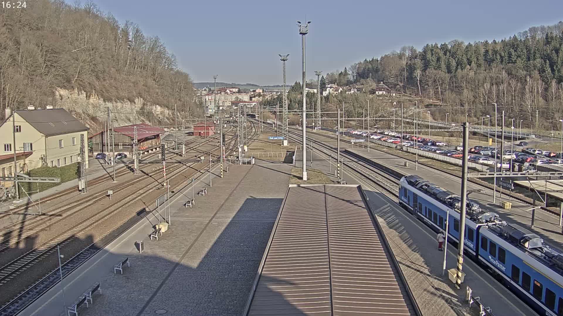 A train station on a sunny day with a blue passenger train at the platform, multiple train tracks, and a large parking lot filled with cars.