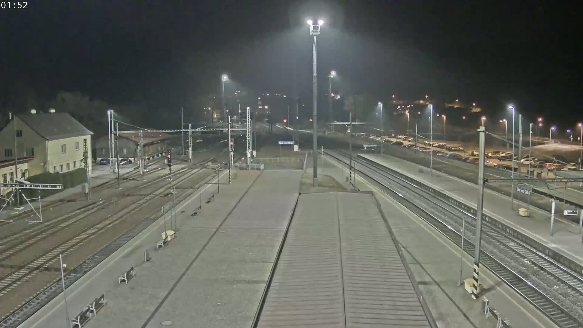 A nighttime view of a train station with multiple train tracks, platforms, and a parking lot, illuminated by artificial lights.