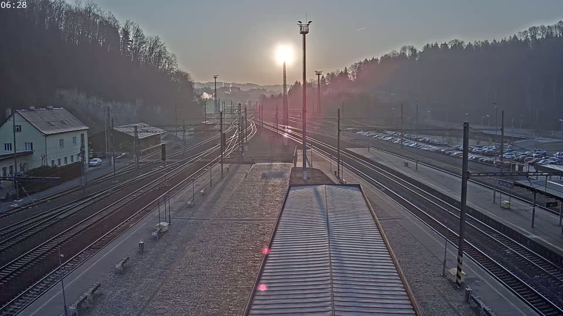A sunlit train station with multiple train tracks, a parking lot filled with cars, and a building in the background on a hazy morning.