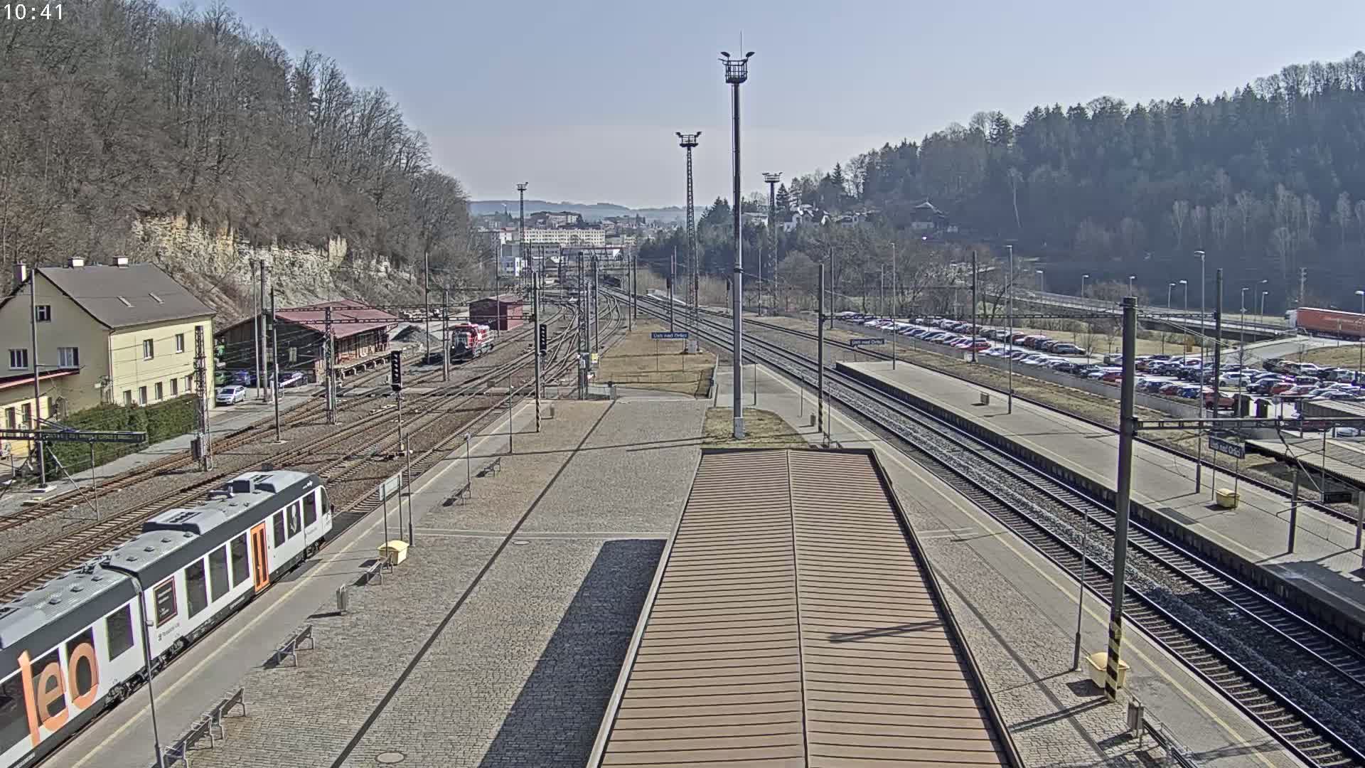 A train station on a sunny day, with multiple train tracks, a passenger train, and a large parking lot filled with cars.