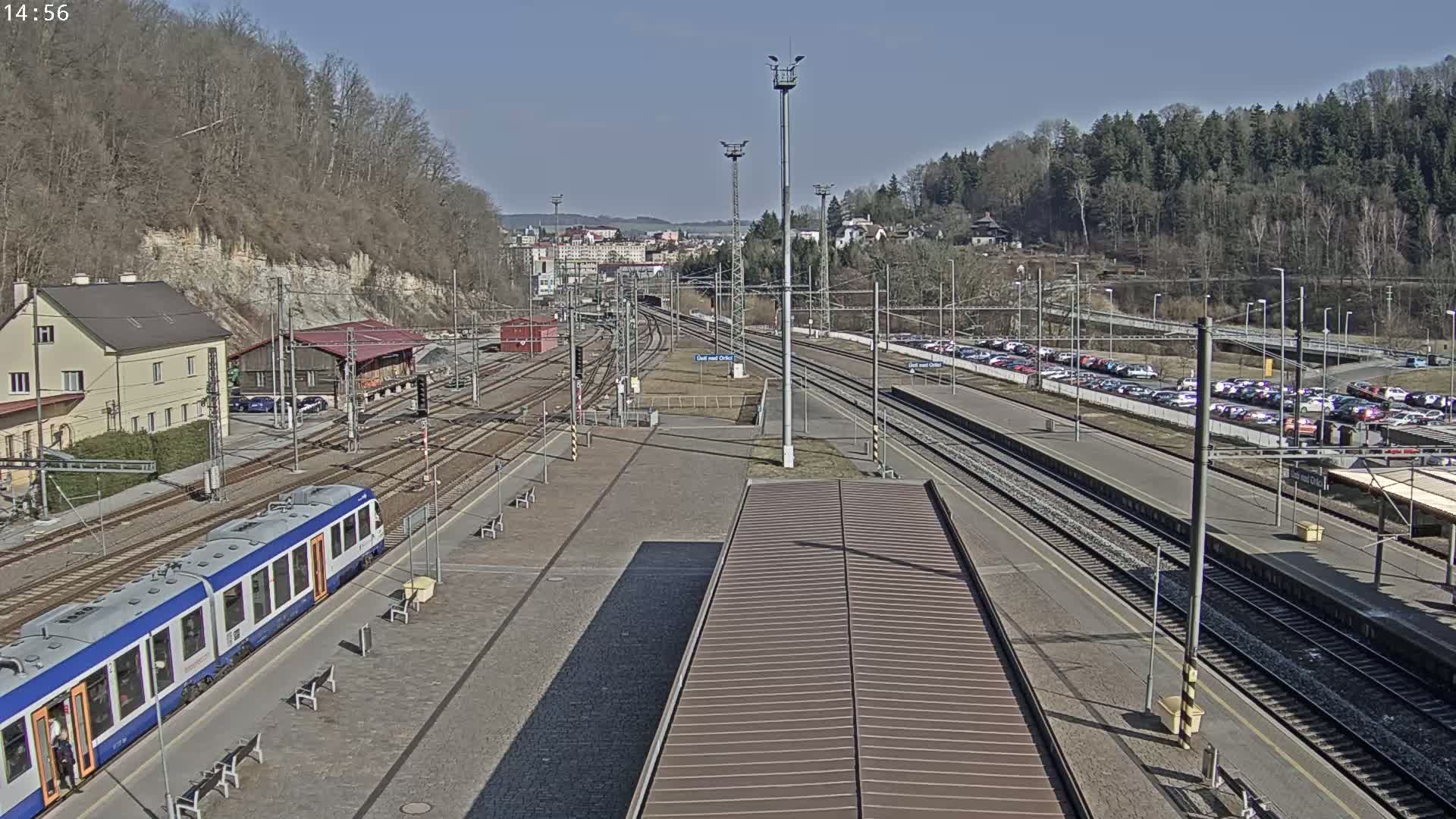A train station on a sunny day with a train at the platform, multiple train tracks, and a large parking lot.