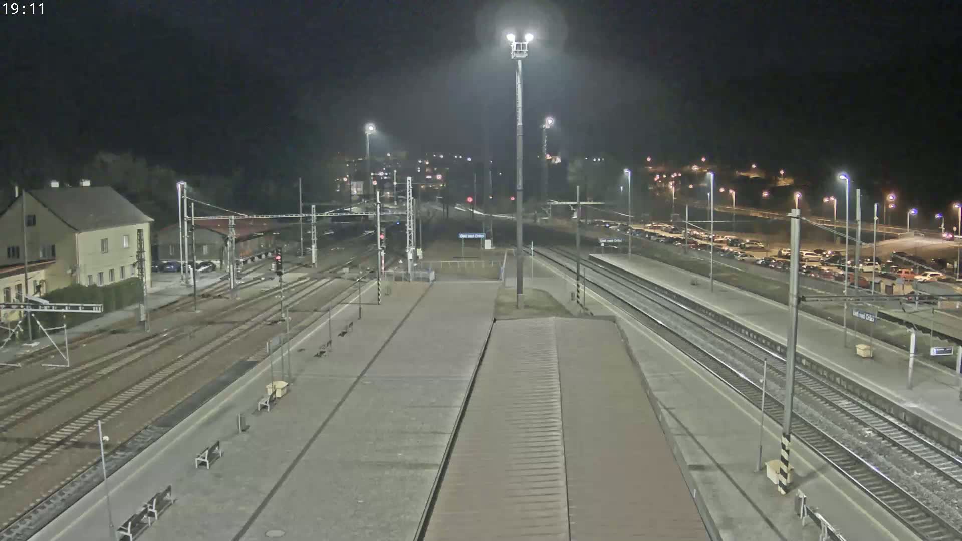 A nighttime view of a train station with multiple tracks, platforms, and a parking lot filled with cars under well-lit conditions.