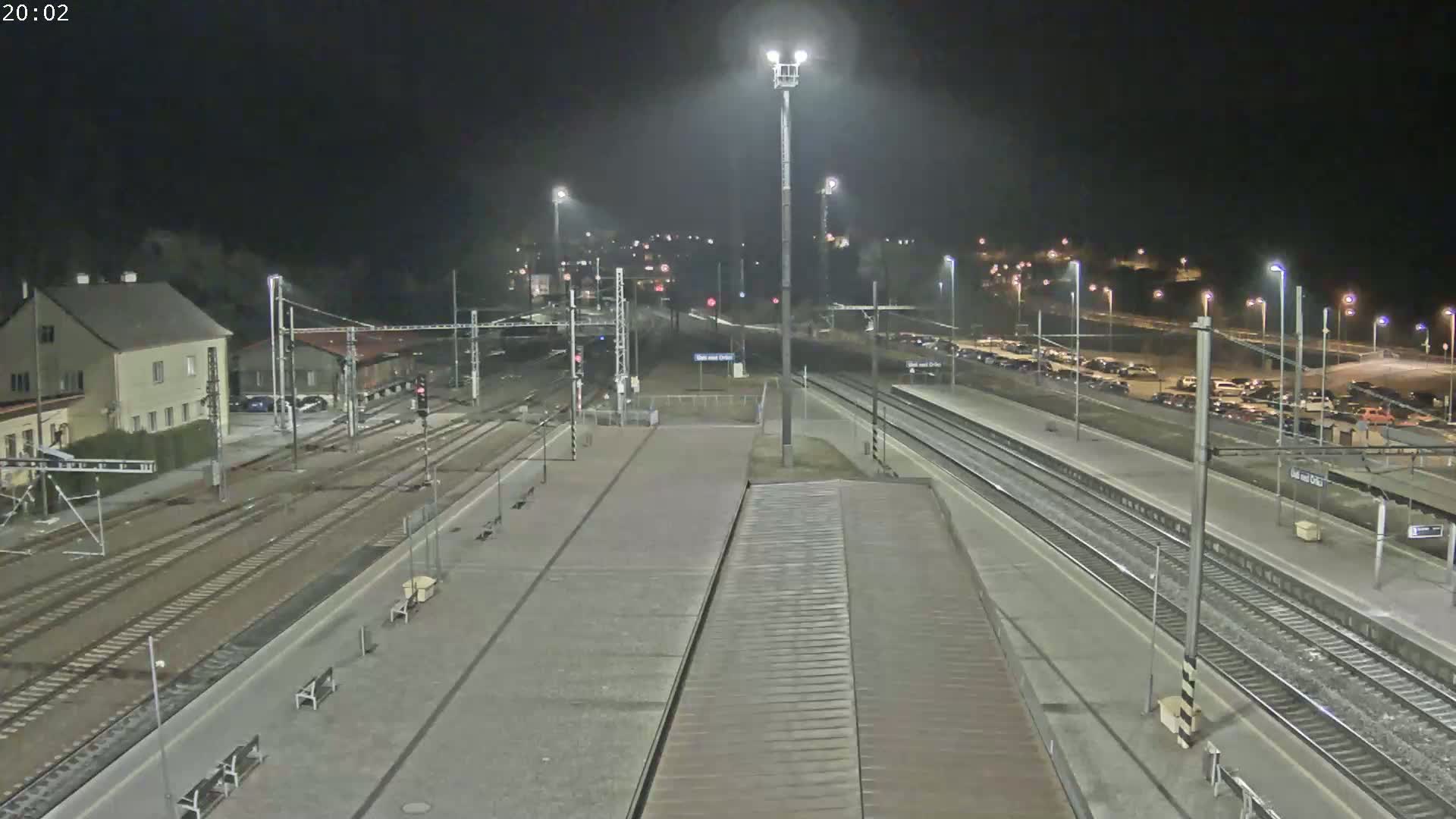A nighttime, high-angle view of a train station with multiple train tracks, platforms, and a parking lot filled with cars.