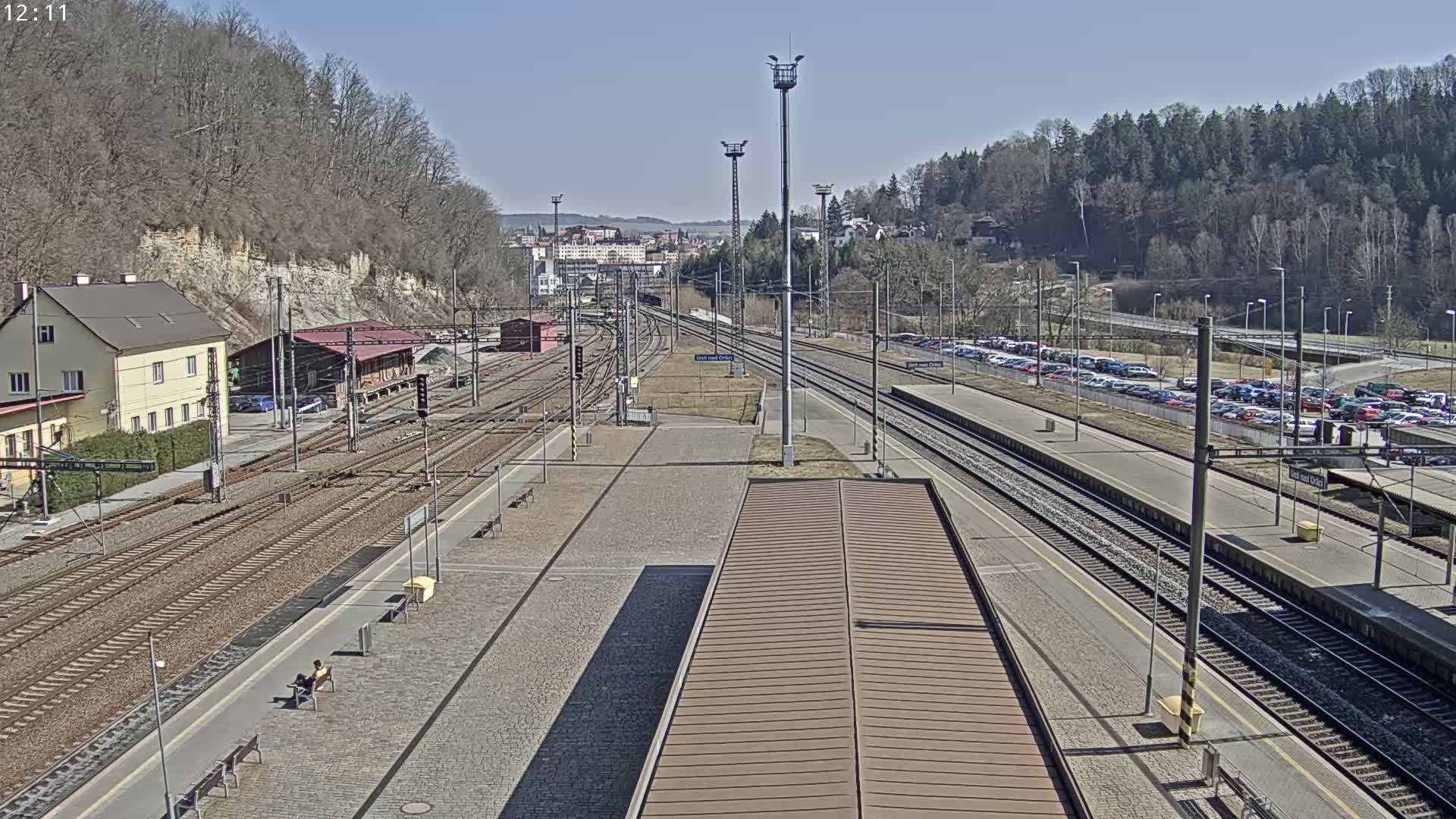 A sunny day at a train station with multiple tracks, a building, and a large parking lot filled with cars.