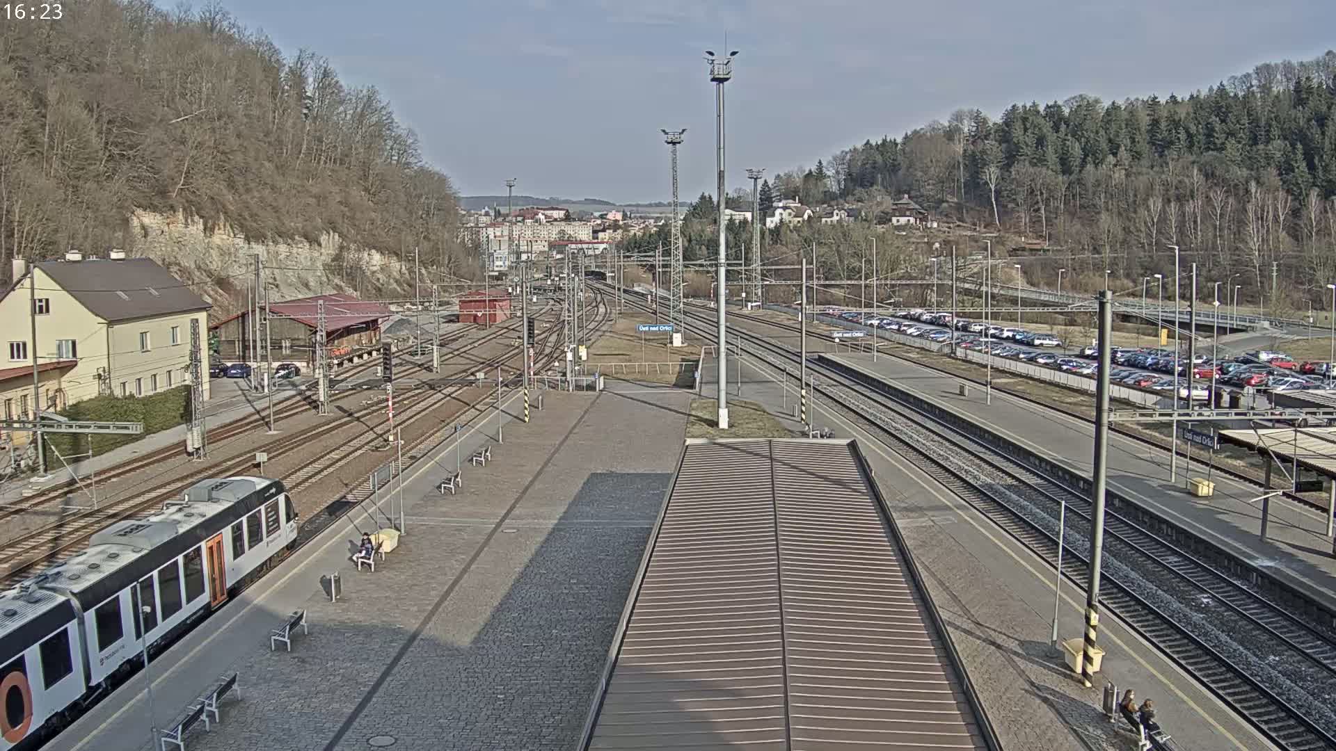 A train station on a sunny day with several train tracks, a partially visible train, a large parking lot, and a wooded hillside in the background.