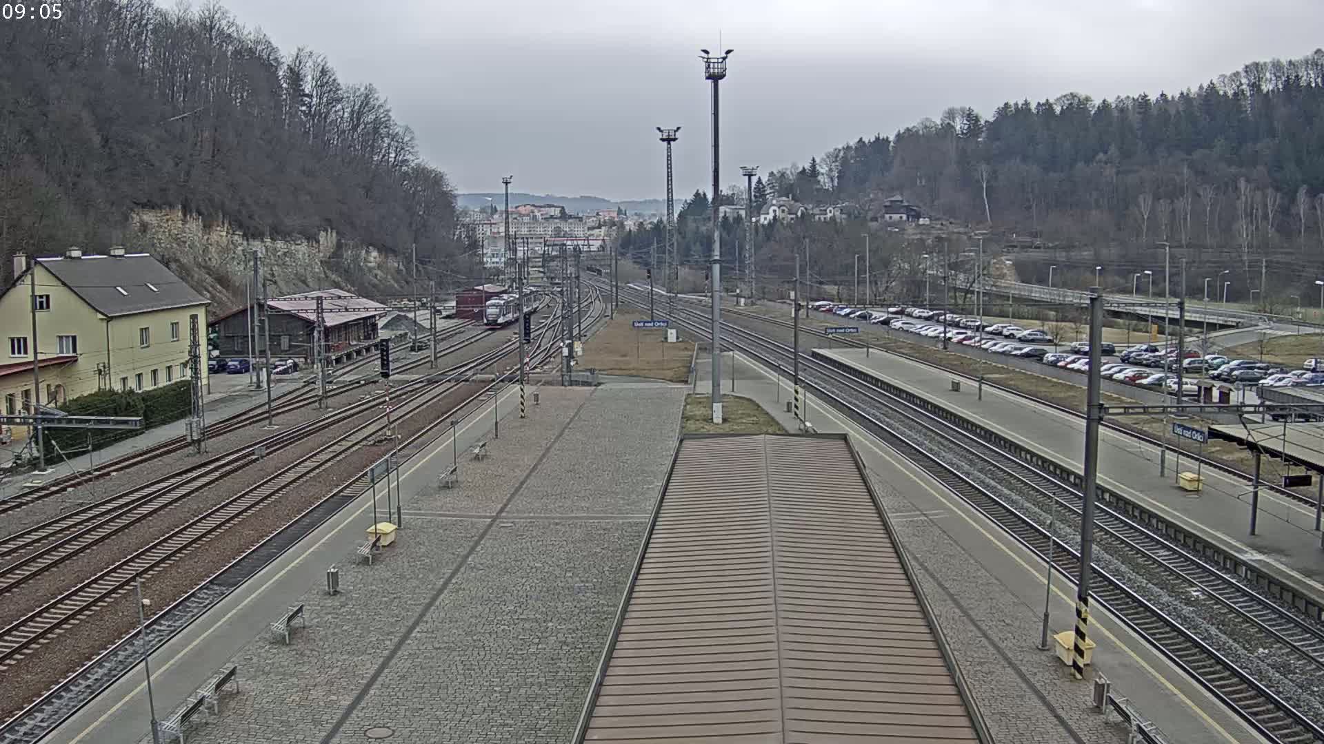 A train station with multiple tracks, a train, a parking lot full of cars, and buildings, all under an overcast sky.