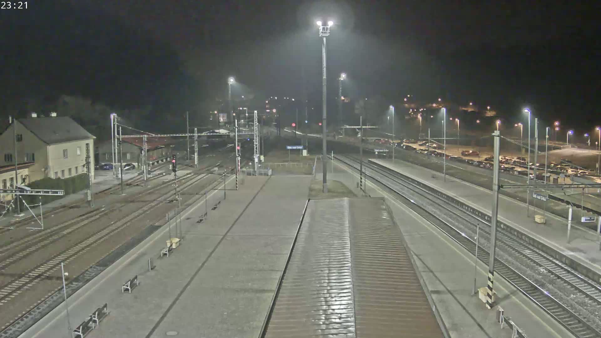 A nighttime view of a train station with multiple tracks, platforms, and a parking lot, under slightly overcast conditions.