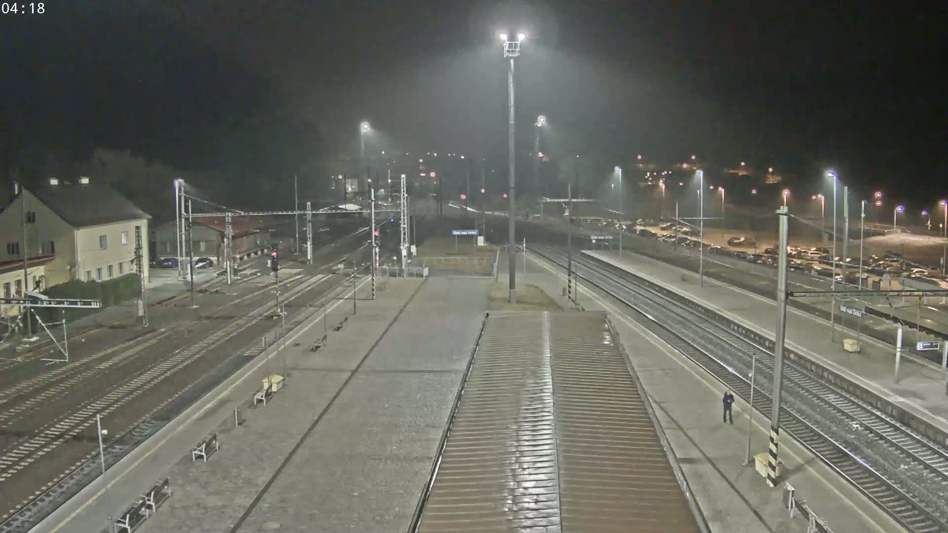 A nighttime view of a train station with multiple tracks, platforms, and a large parking lot, under misty or rainy conditions.