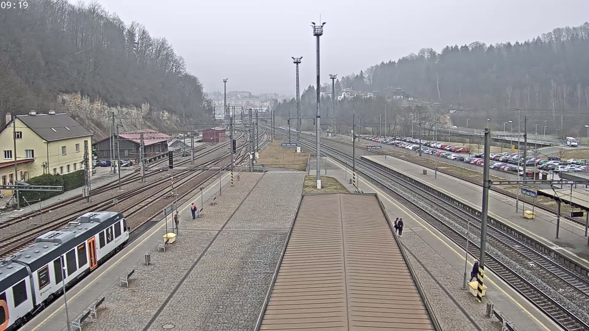 A train station with multiple train tracks, a train at a platform, and a large parking lot, all under an overcast sky.