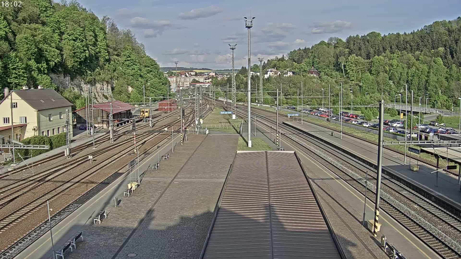 A train station with multiple tracks, a building, and cars parked in a lot on a partly sunny day.