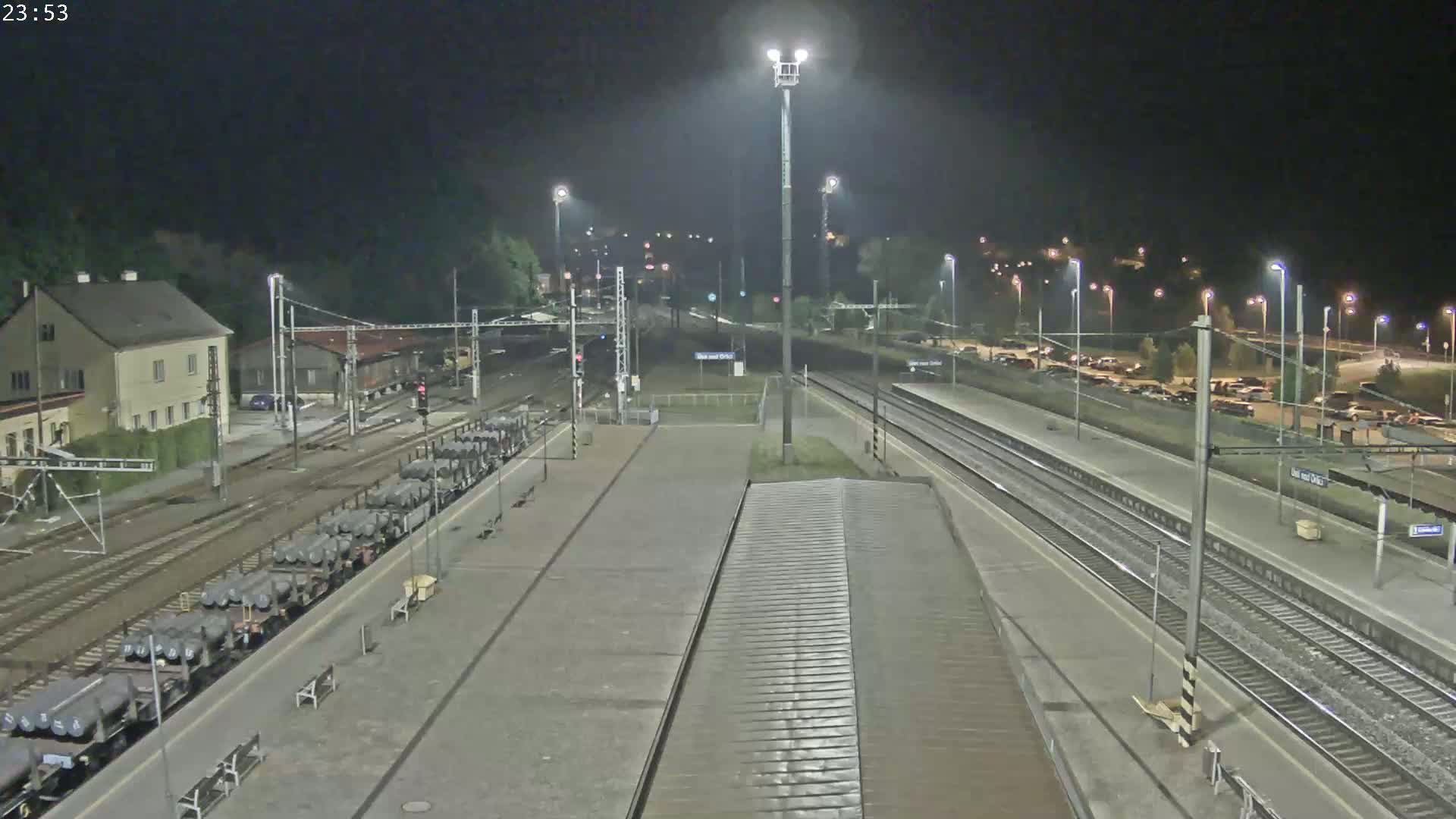 A nighttime view of a train station with several railway tracks, a train carrying cylindrical cargo, and a parking lot with cars.