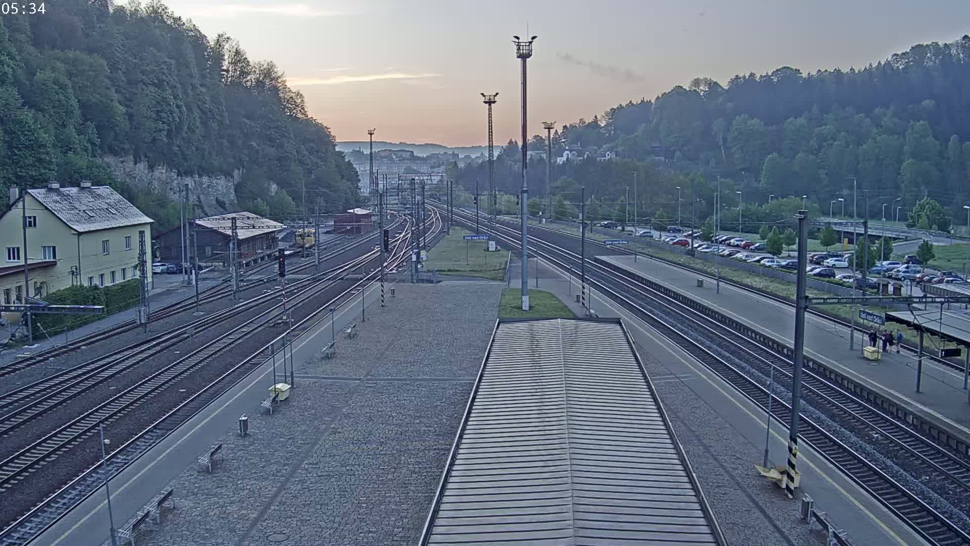 A high-angle, long shot of a train station at dawn, featuring multiple train tracks, a parking lot, and a building, under a mostly clear sky.