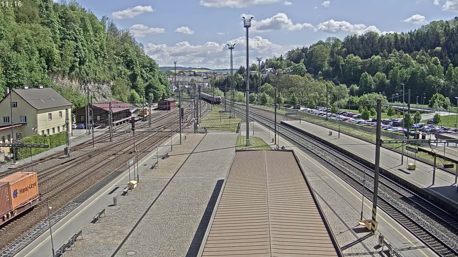 A train station with multiple tracks, a train in the distance, and a partly sunny sky is seen from an elevated vantage point.
