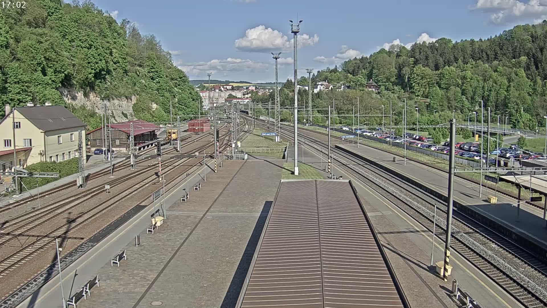A train station with multiple tracks, a parking lot, and buildings surrounded by green hills under a partly sunny sky.