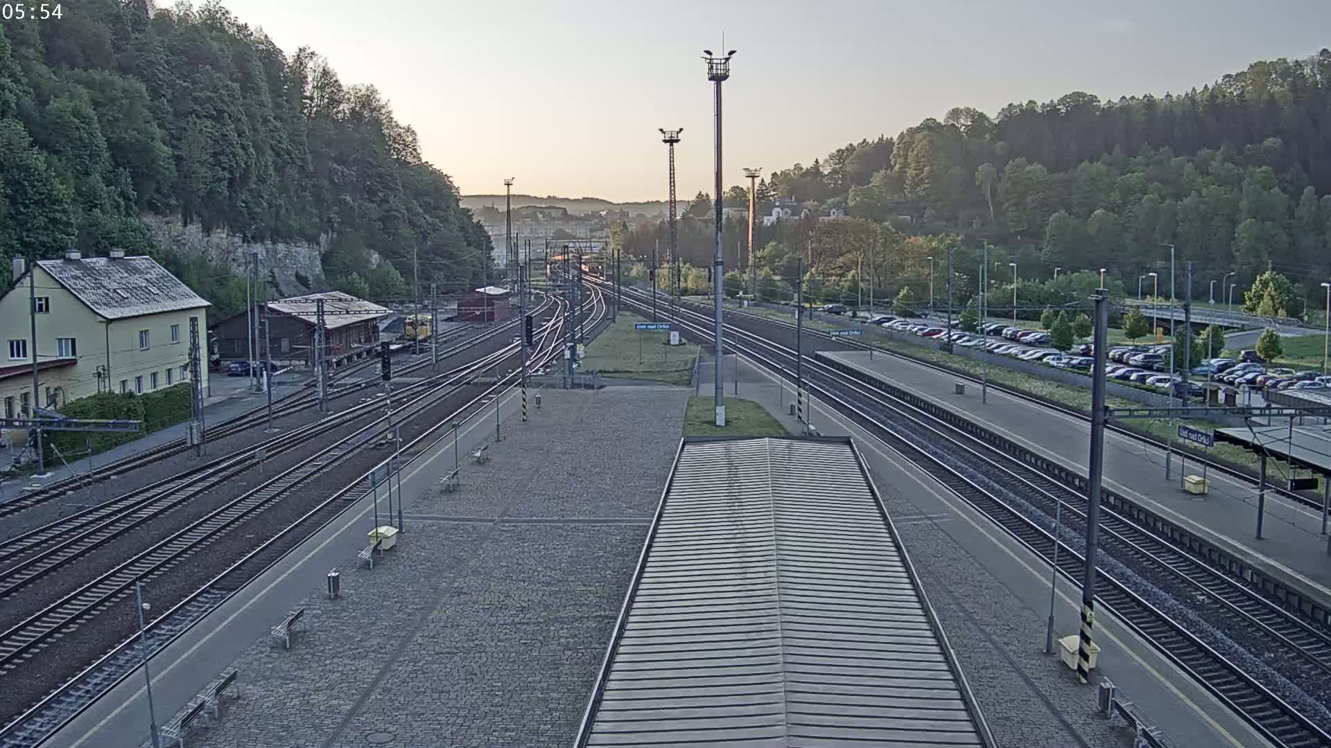 A train station with multiple tracks, a building, a parking lot, and a hillside covered in trees is seen at dawn under a clear sky.