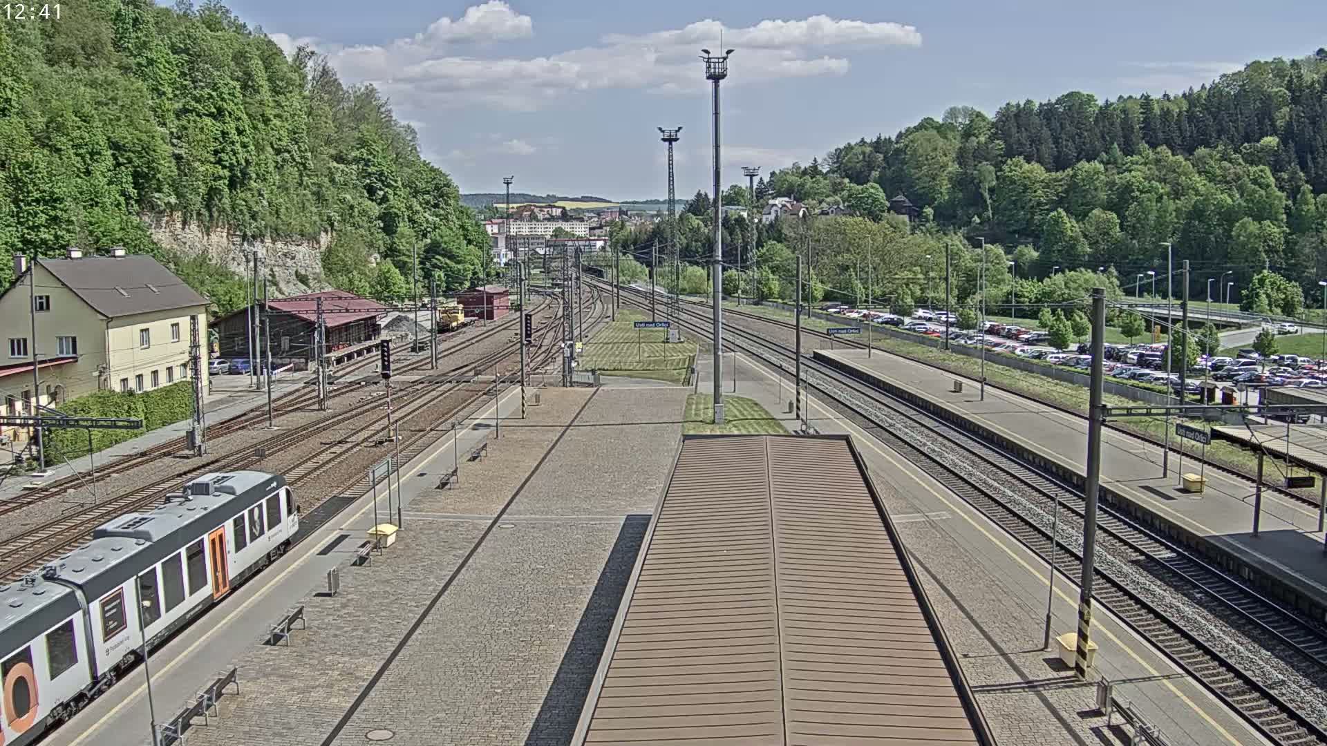 A train station with multiple tracks, a passenger train, and a large parking lot is visible on a sunny day.